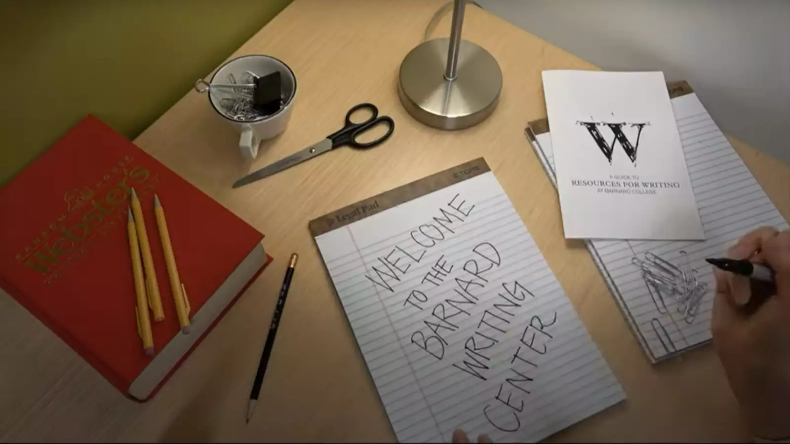 A dimly lit desk with a notepad, pencils and dictionary scattered across it.